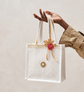 Hand holding a decorative tote bag with a red flower and gold bow on a light background
