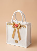 Decorative white tote bag with a gold bow and pink flower on a beige background